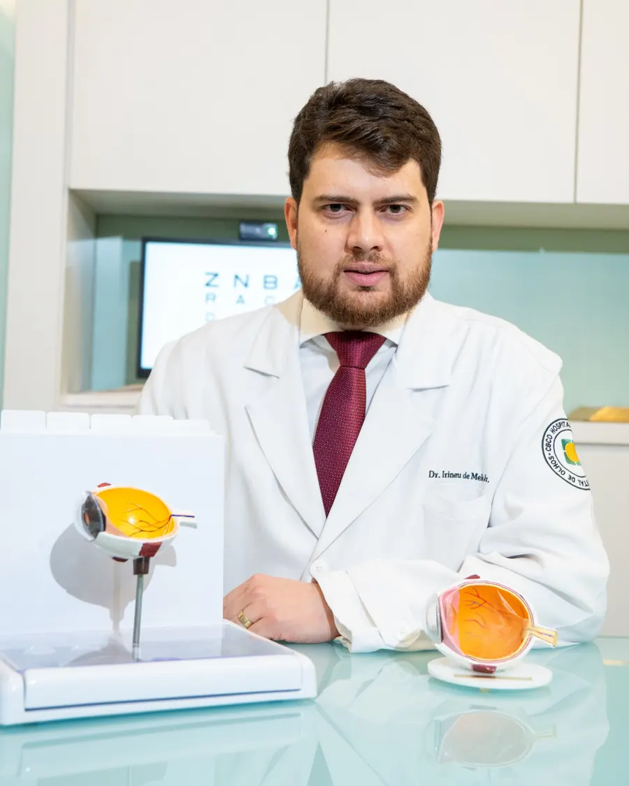 Dr. Irineu à mesa do consultório, com modelos anatômicos do olho usados para explicar o diagnóstico